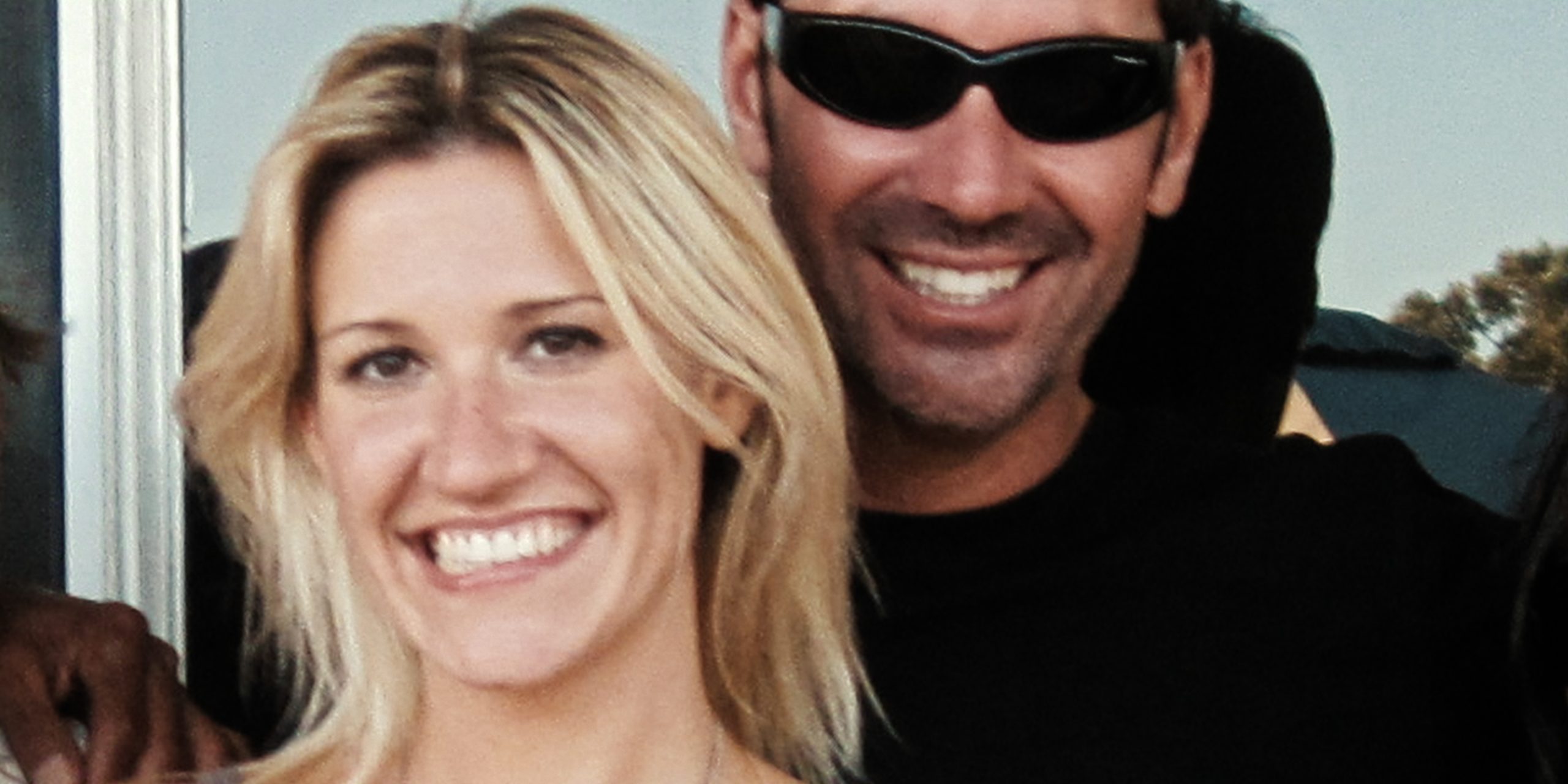 "Smiling woman with blonde hair and a man in sunglasses standing close together outdoors, both appearing happy with sunlight and greenery in the background."