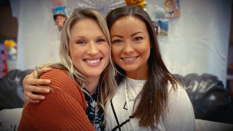 "Two women smiling and sitting close together on a black couch, embracing each other in a cozy indoor setting with photos and decorations in the background."