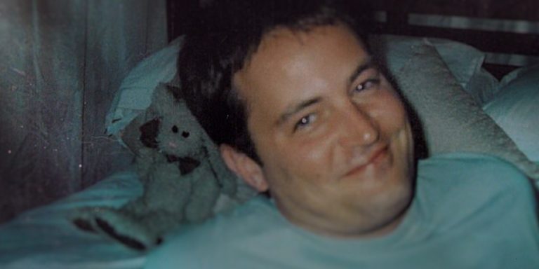 "Smiling man lying on a bed with white pillows and a teddy bear in the background, in a softly lit bedroom environment."